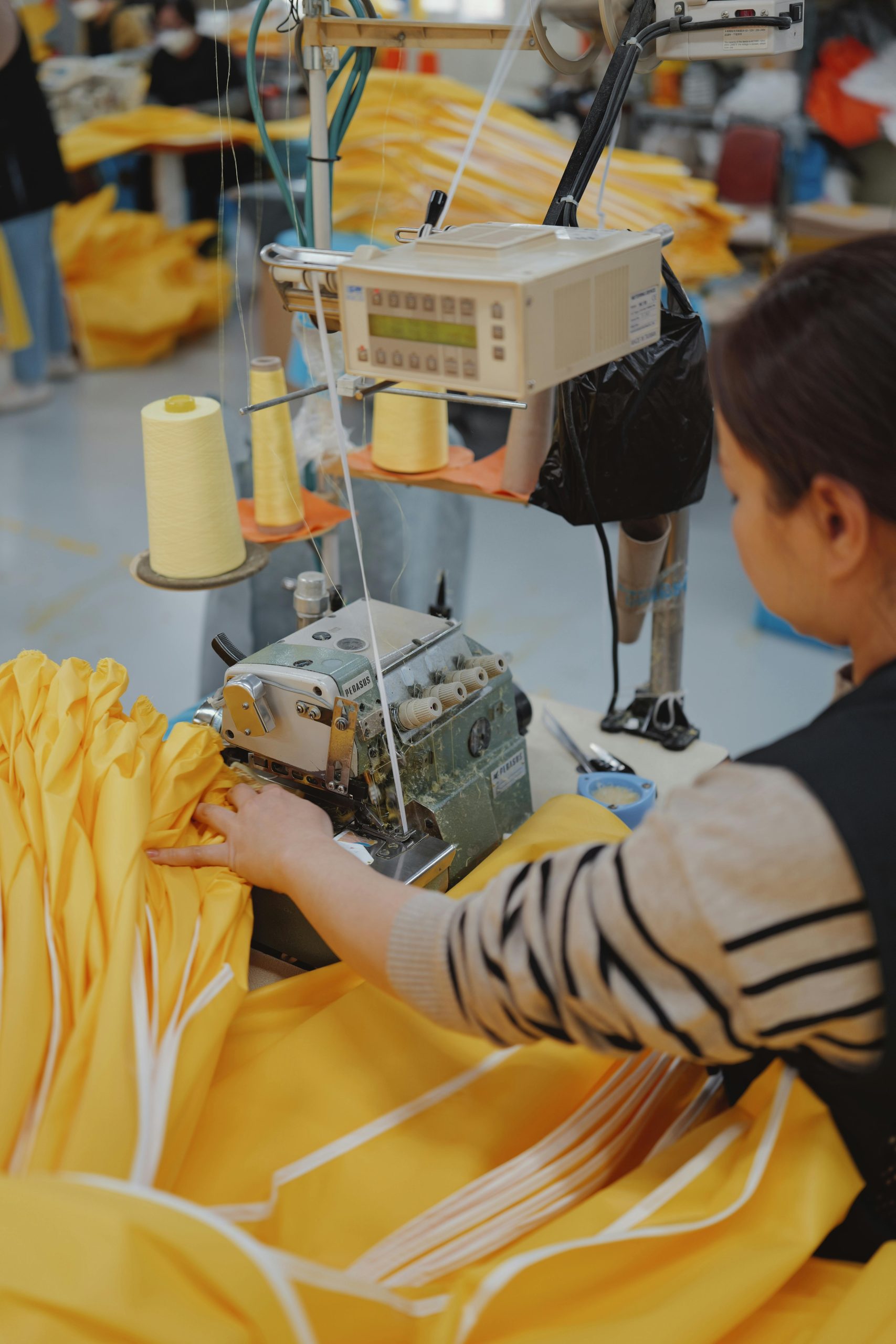 Textile worker using a sewing machine to stitch yellow fabric in a factory setting.